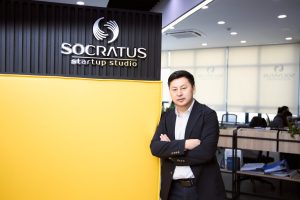 A person in a suit stands with arms crossed in front of a yellow and black wall with the Socratus Startup Studio logo. The background features a modern office space with desks and chairs.