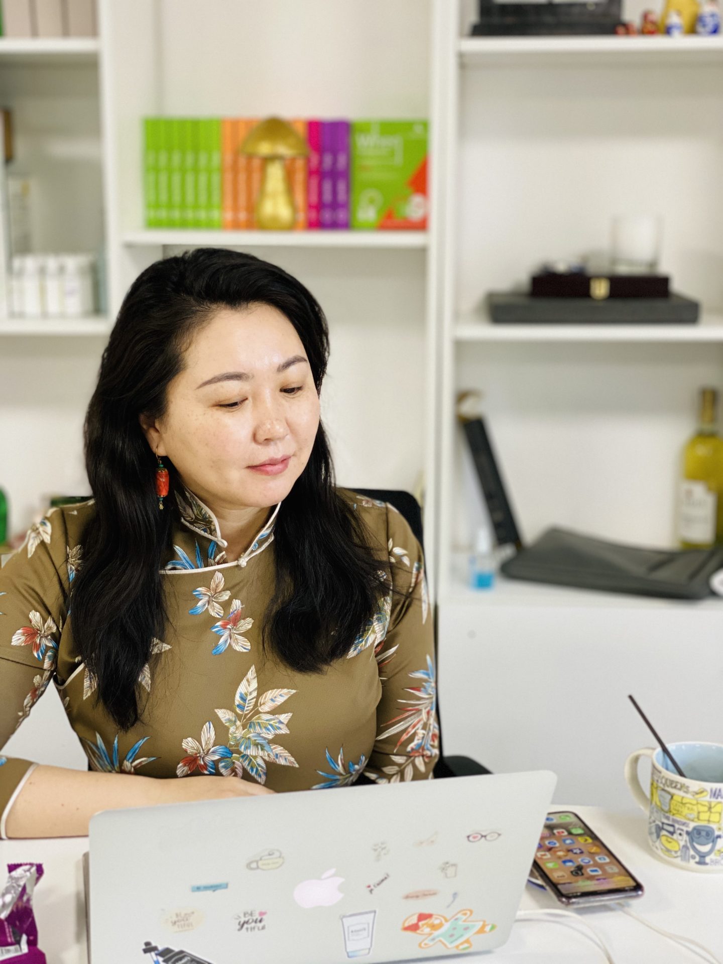 A woman with long dark hair sits at a desk with a laptop. She wears a floral dress and red earrings. The desk has a smartphone, a mug, and stationery. Books and decor items are visible on the shelves behind her.