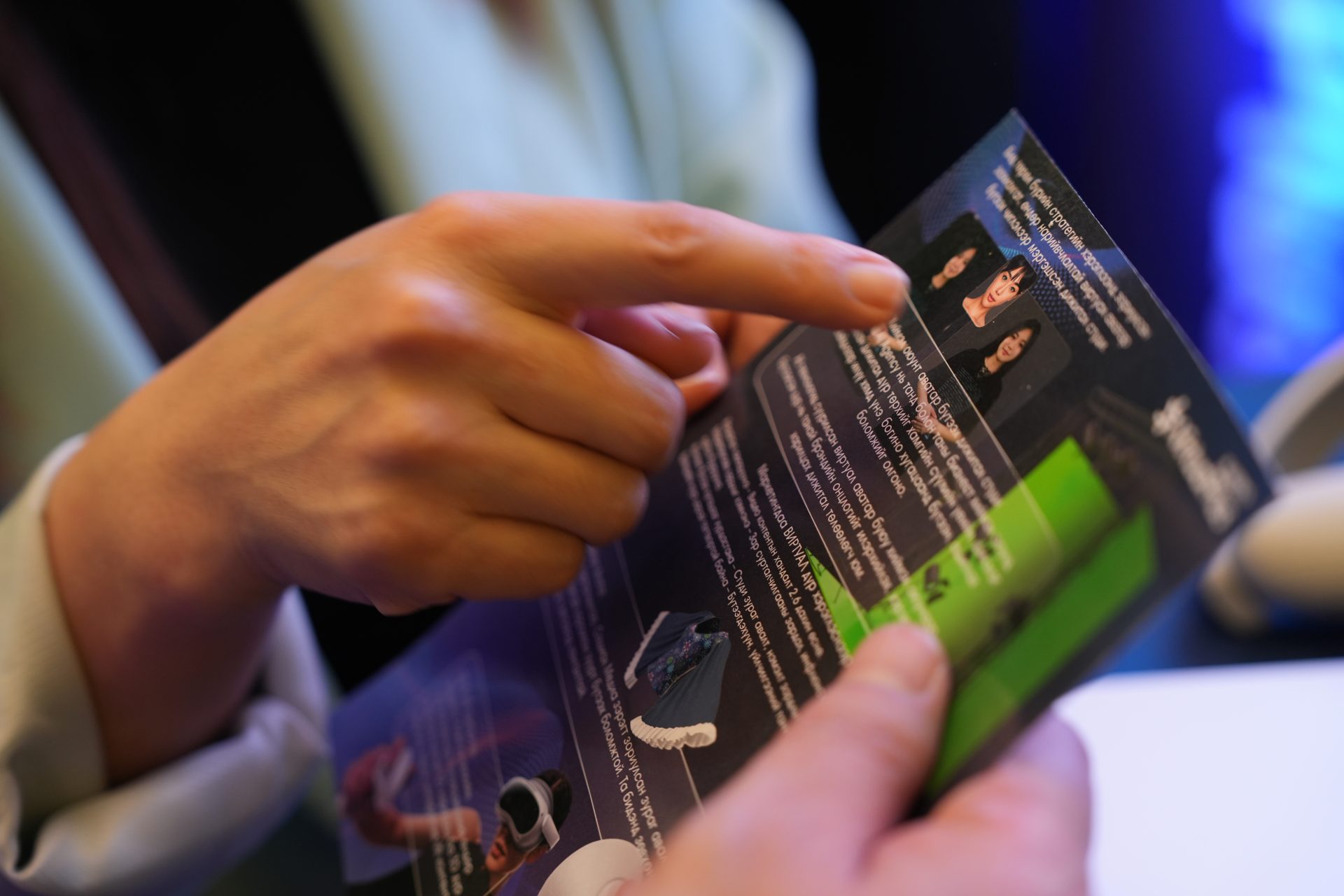 Person holding and pointing at a brochure with text and images. The brochure appears to be informative, possibly detailing products or services. The person's hands are visible, and the background is blurred, emphasizing the focus on the brochure.