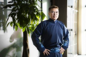 A person wearing glasses and a dark blue traditional outfit with embroidered designs stands confidently with hands on hips. They are indoors next to a large plant, and sunlight streams through nearby windows.