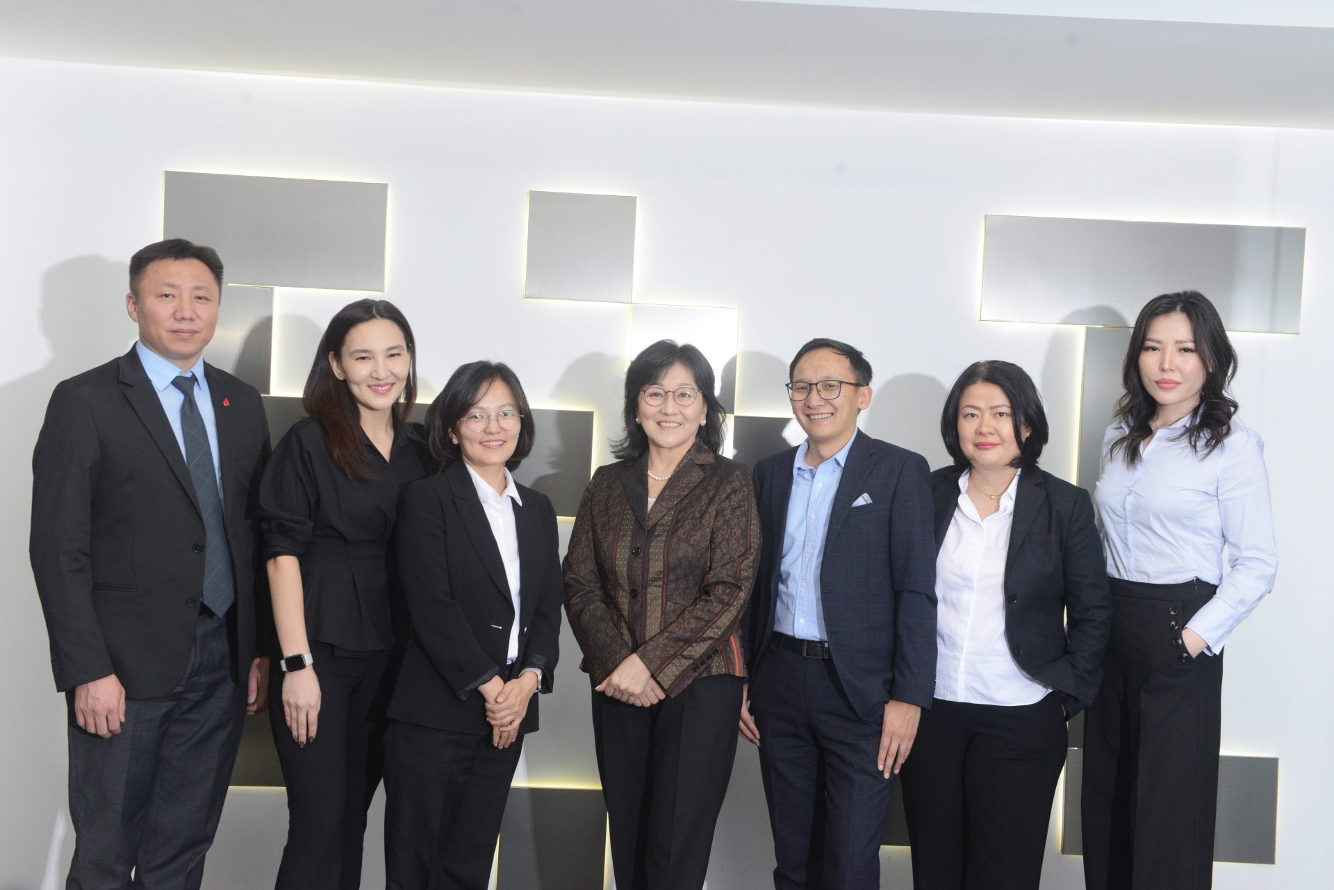 Seven people in formal attire standing side by side, posing for a photo against a white wall with geometric patterns.