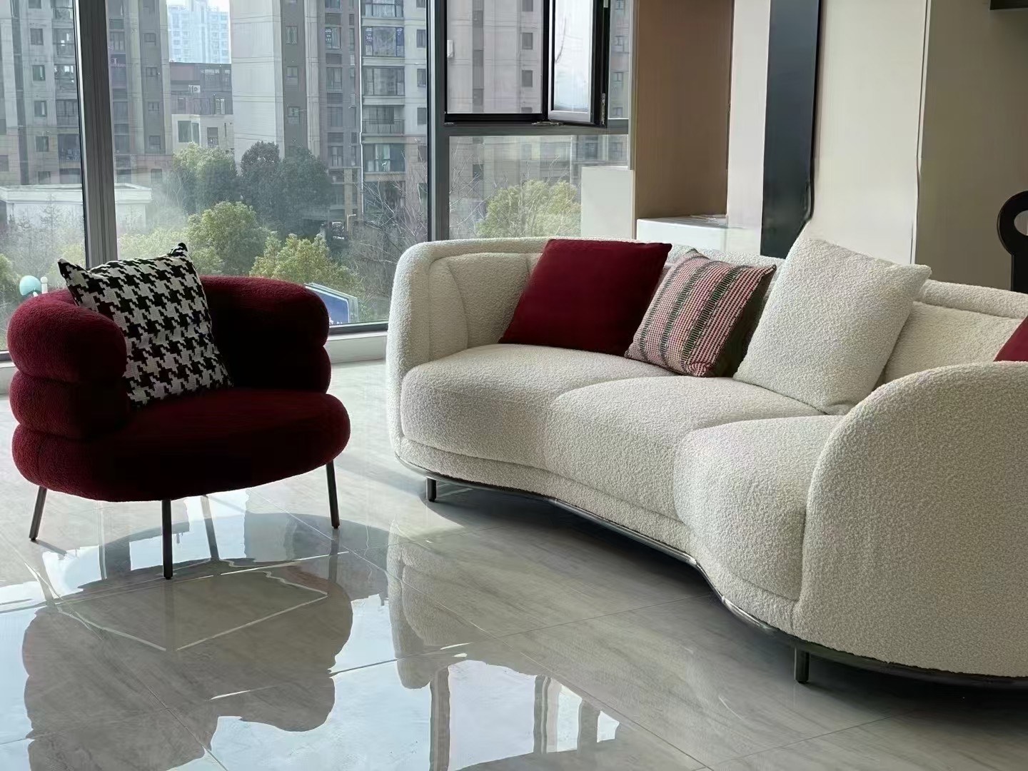 Modern living room with a large curved white sofa and red cushions, paired with a red armchair featuring a black-and-white houndstooth cushion. The space has large windows overlooking city buildings, and the shiny floor reflects the furniture.