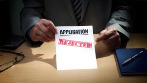 A person in a suit sits at a desk holding a paper labeled "APPLICATION" with a large red stamp reading "REJECTED." The desk also has a notebook and a computer keyboard. The room is dimly lit, highlighting the document.