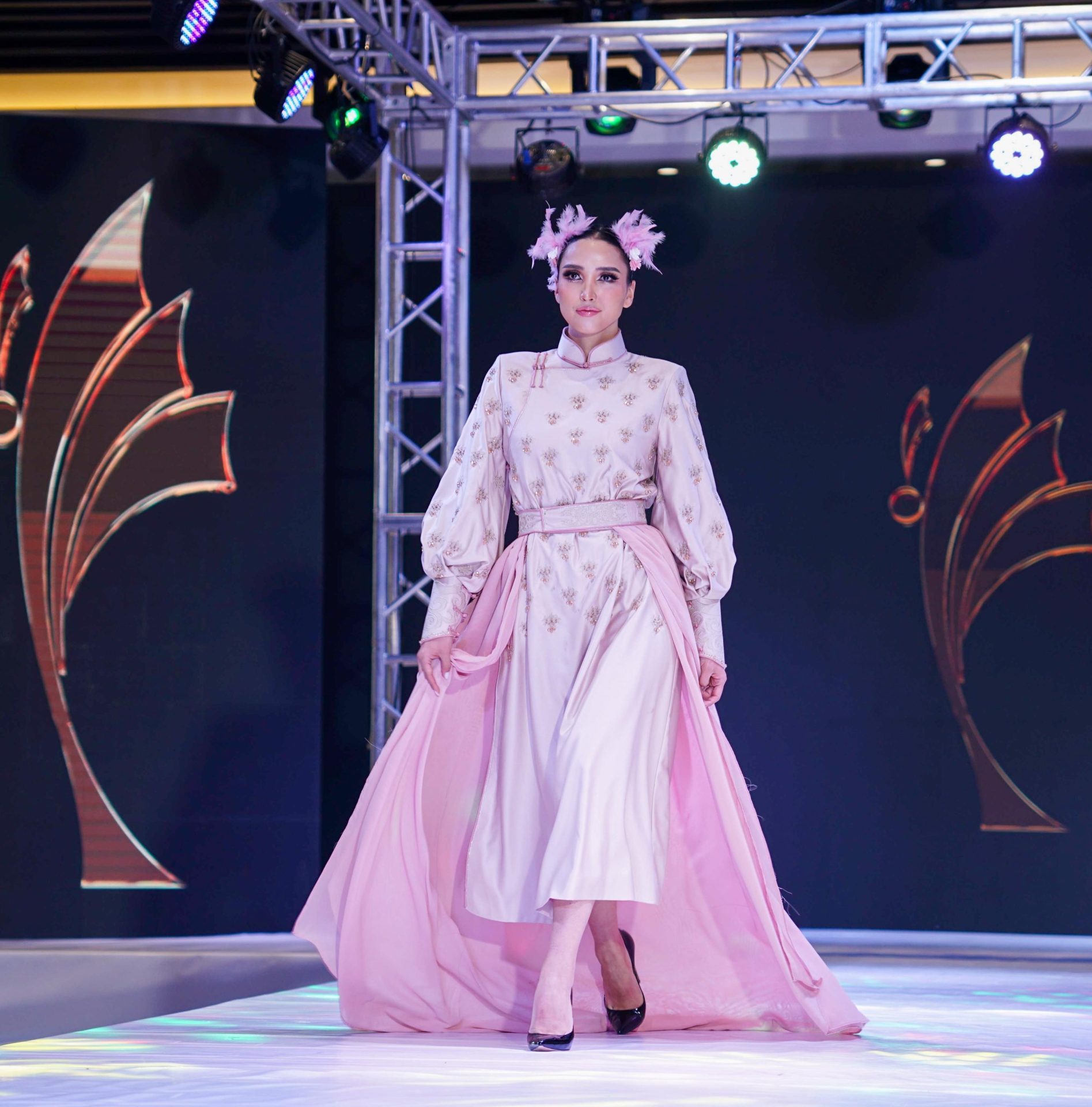 A model walks down the runway wearing a long-sleeved, high-collared dress with delicate patterns and a flowing pink skirt. Her hair is adorned with pink flowers. The stage features elegant abstract designs and overhead lighting.