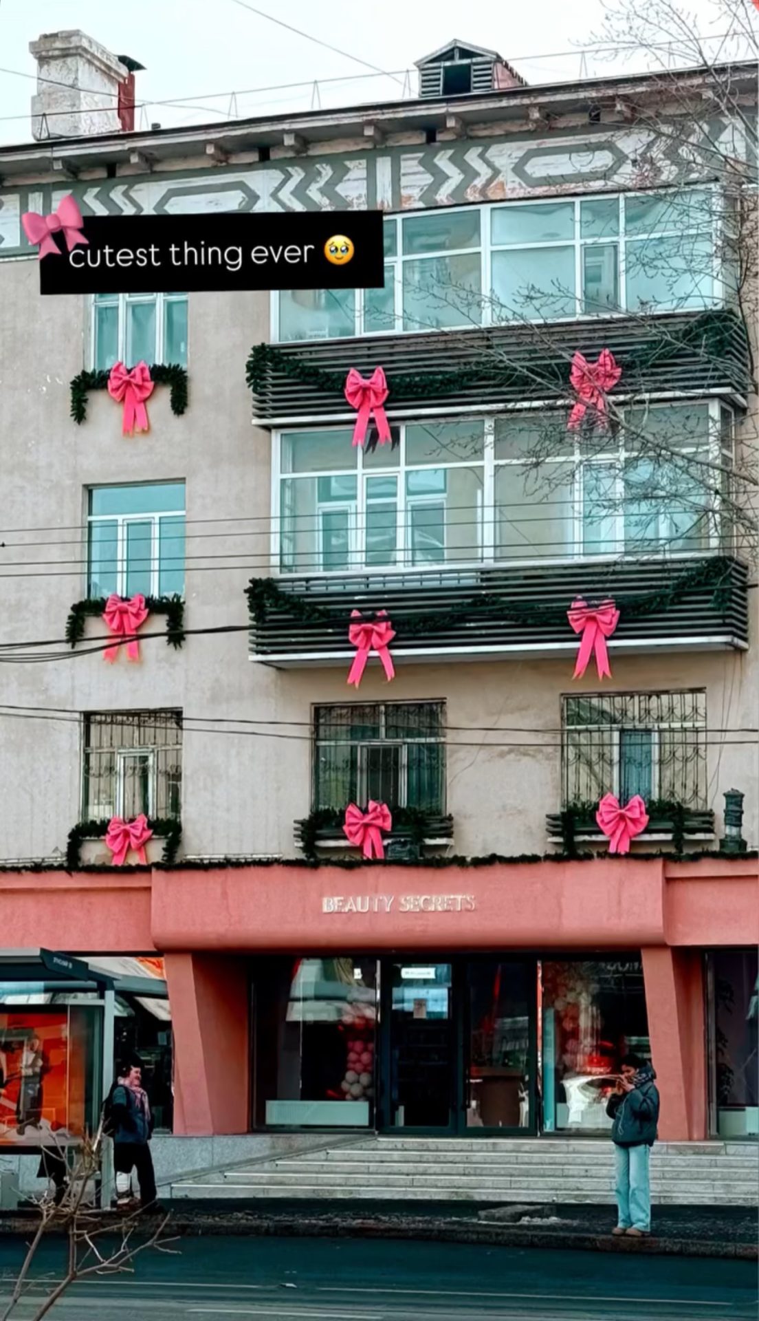 A building with large pink bows and green garlands decorating the windows. The sign says "Beauty Secrets." Two people stand outside. A text overlay in the image reads "cutest thing ever" with a happy face emoji.