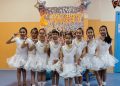 Seven young girls in matching white dresses stand in a line, posing with peace signs. They are in front of a sparkling backdrop with "SMART" in neon lights and balloon decorations.