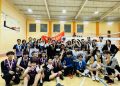 A large group of young people, some wearing sports uniforms and holding medals, pose for a group photo in an indoor sports hall. A basketball hoop is visible, and a bright orange banner is in the background.