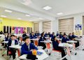 A classroom filled with students sitting at desks, wearing blue sweaters and white shirts. The classroom has bright walls with decorations and a whiteboard. The students are facing forward, with smiles and attentive expressions.