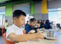 A group of children sitting at a table in a brightly colored room. A boy in the foreground focuses on preparing a sandwich. Other children are engaged in similar activities in the background, with trays and cups on the table.