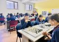 Students in a classroom are engaged in playing chess. They sit at tables, concentrating on their games. A sign with the number 25 is displayed on a table. In the background, a teacher oversees the activity. The room has yellow walls and windows.