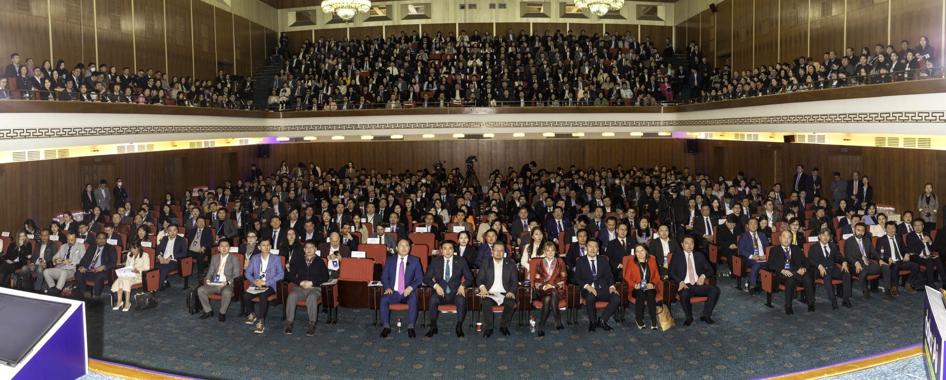 A large group of people seated in a spacious auditorium, facing the front. The space is decorated with wood paneling, chandeliers, and numerous rows of chairs filled with men and women, all dressed in formal attire.