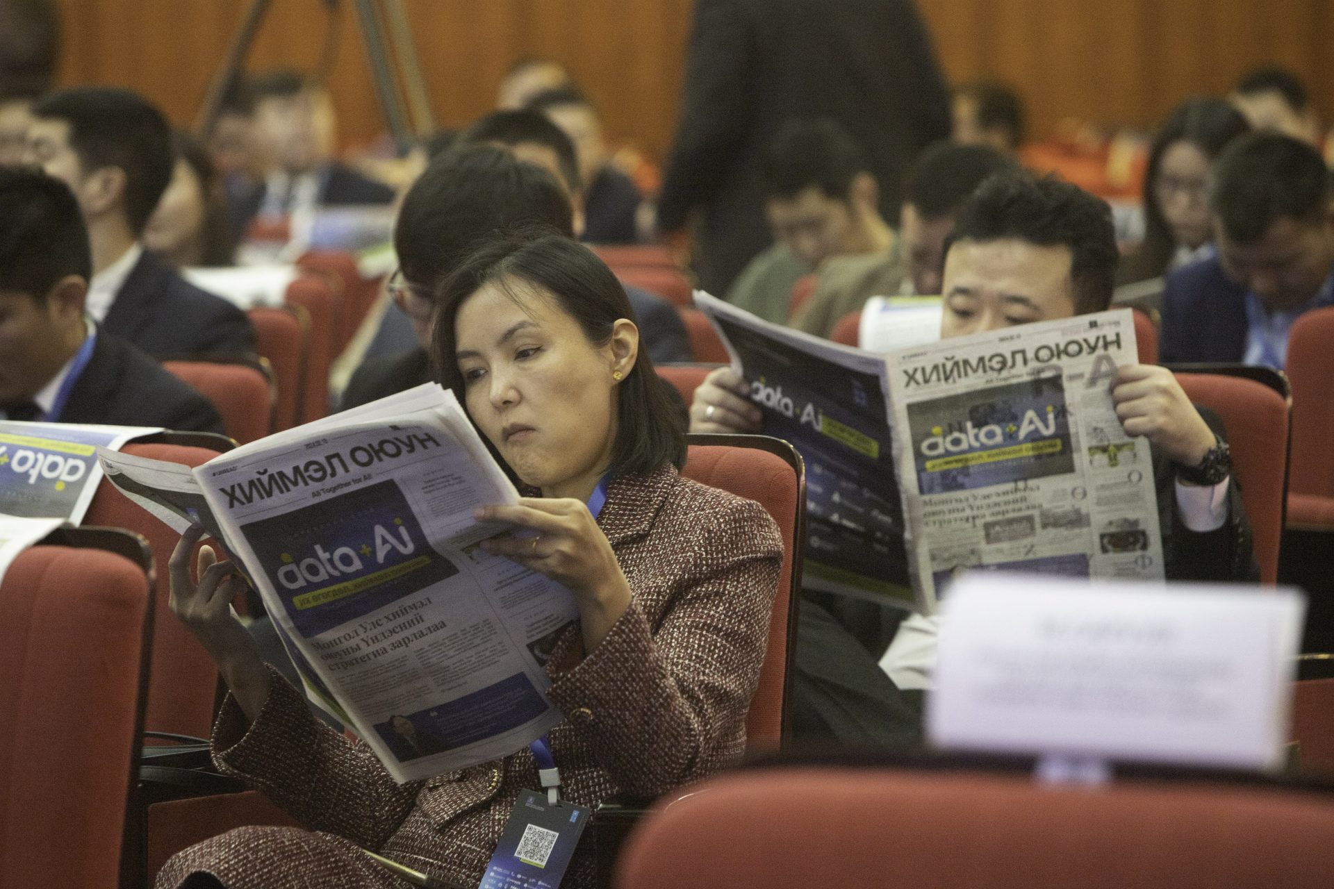 People seated in rows, reading newspapers at an indoor event. They are dressed formally, suggesting a professional or business setting. The newspapers have a headline in Cyrillic script.