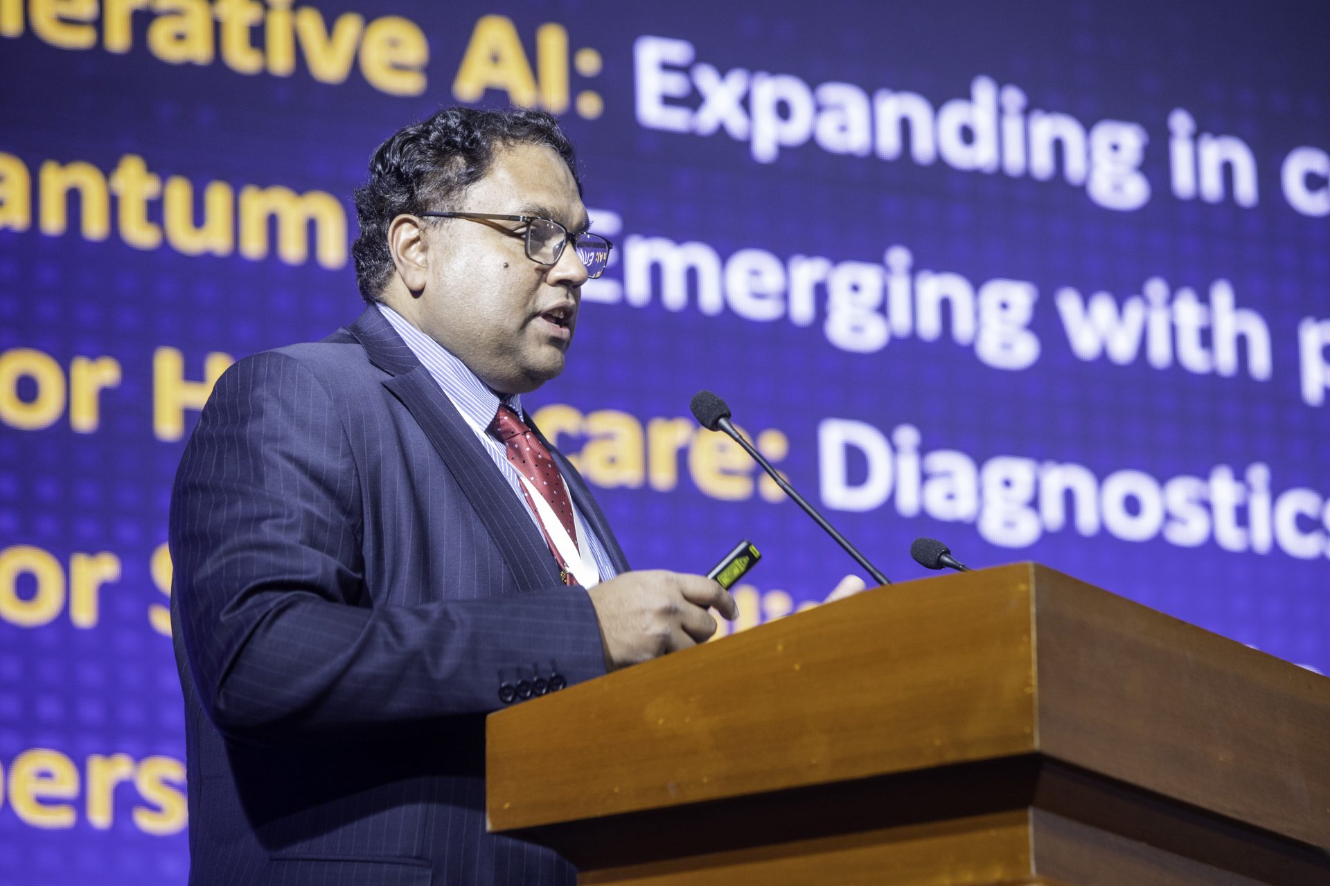 A person in a suit speaks at a podium with a microphone, holding a smartphone. Behind them is a large screen displaying text related to AI and healthcare topics.