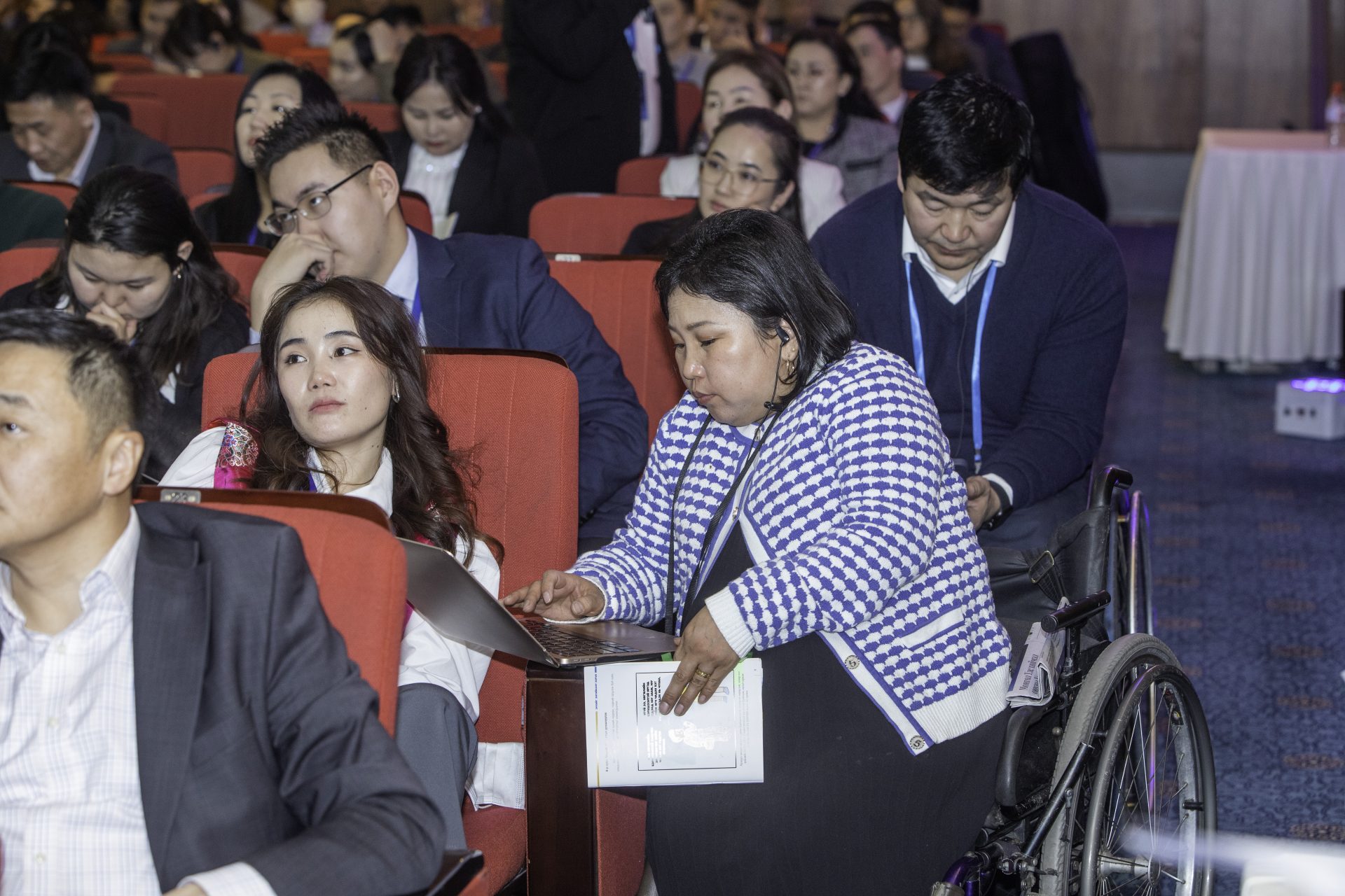 A group of people sitting in a conference hall. A woman in a wheelchair works on a laptop, while others nearby listen attentively. Many attendees appear engaged, seated in rows of red chairs.