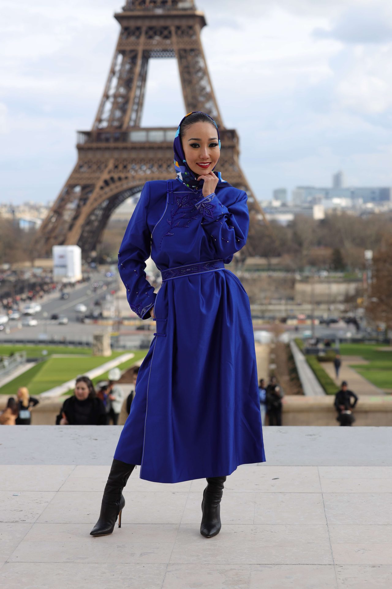 A woman in a vibrant blue dress and matching headscarf poses confidently in front of the Eiffel Tower. She stands on a paved area, with people and a cityscape in the background. She wears black boots and touches her chin with one hand.