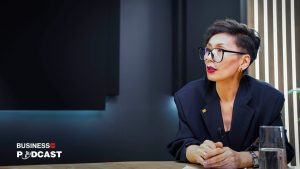 Person wearing large glasses and a black blazer sits at a table in a modern studio setting, with the words "Business Podcast" visible on the lower left. Bright vertical lights and dark paneling serve as the background.