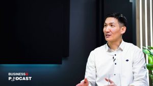 A man in a white shirt sits and gestures with his hands, speaking in front of a dark background. A logo on the wall says "Business Podcast." Vertical stripes and a plant are partially visible on the side.