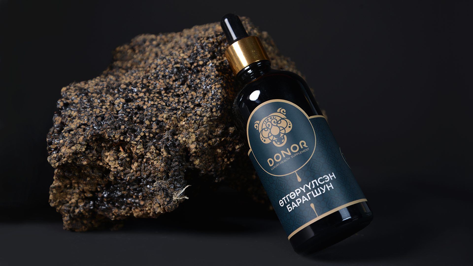 A dark bottle labeled "DONOR" with intricate design rests against a large, textured rock on a black background. The bottle has a dropper cap and gold accents.