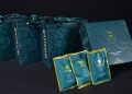 Teal gift bags and a matching box with gold ornate designs are displayed. Two packets with the same design are in front. The bags have ribbon handles, and the box features a stylized emblem. The overall theme is elegant and luxurious.