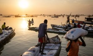 People on boats transport large sacks during a sunset on a calm body of water. The sky is orange, reflecting on the water's surface. Some individuals carry sacks on their heads while others load them onto boats. Distant trees and boats are silhouetted.