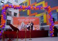 A stage decorated with purple and orange balloons features a red grand piano and a backdrop with illustrations of people and a festive message. The event takes place in front of a colorful building with large windows.