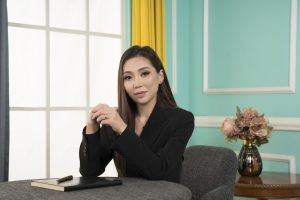 Woman sitting at a desk with hands clasped, wearing a black blazer. A notebook and pen are on the table. Light teal walls, a vase of flowers, and a large window with a yellow curtain are in the background.