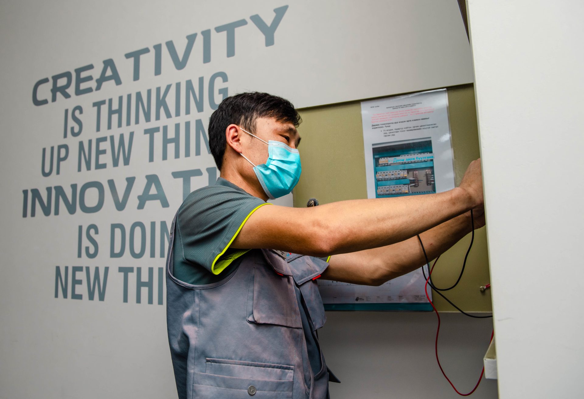 A person wearing a face mask and gray uniform is working on an electrical panel. In the background, there's a motivational wall text about creativity and innovation.