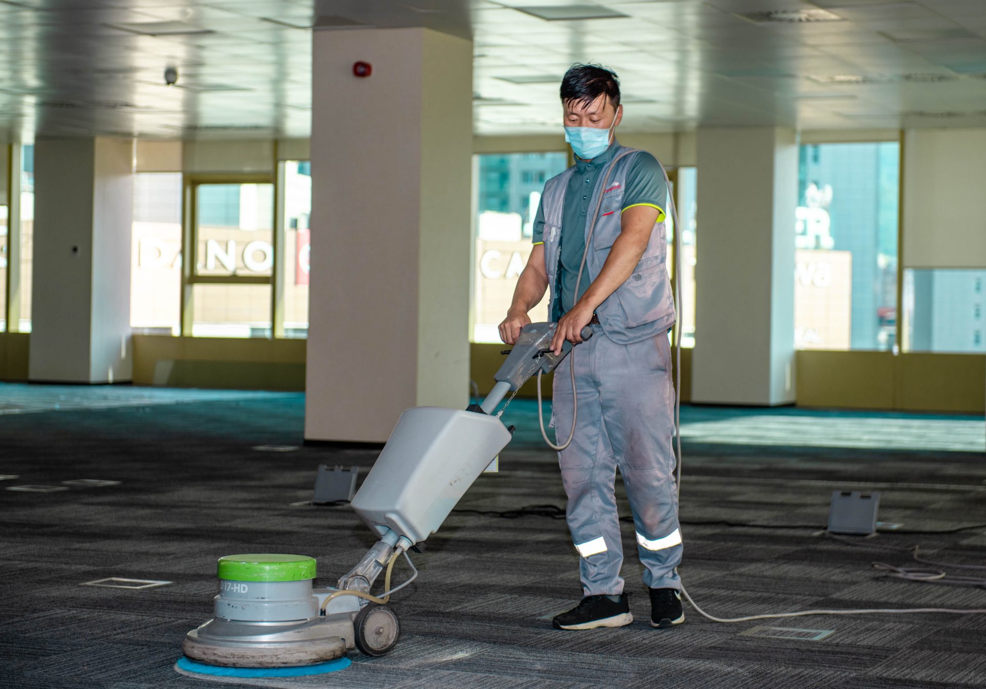 A person wearing a face mask and gray work uniform operates a floor polishing machine in a large, empty room with carpet flooring and large windows.