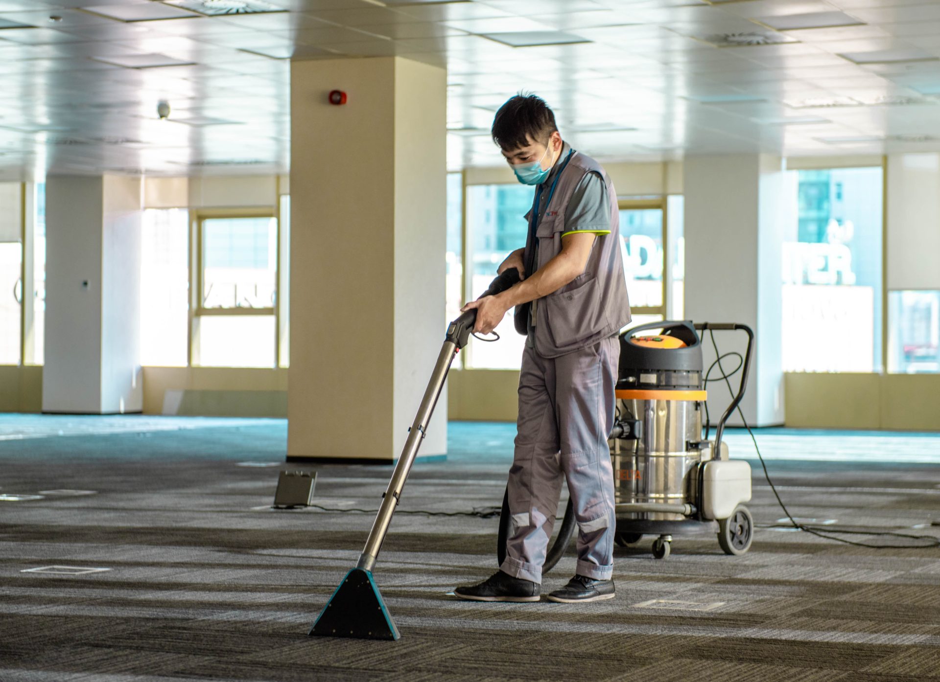 A person wearing a face mask and gray uniform is using a carpet cleaning machine in a large, empty room with tall windows. The machine has a hose and a canister on wheels. Sunlight is filtering through the windows onto the carpet.