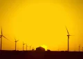 Silhouetted wind turbines and a house are seen against a vibrant orange sunset sky, with the sun sitting low on the horizon.