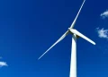 A large white wind turbine stands tall against a vibrant blue sky, with a few small clouds in the distance. The turbine's blades are visible, positioned at an angle, capturing the clear and open atmosphere.