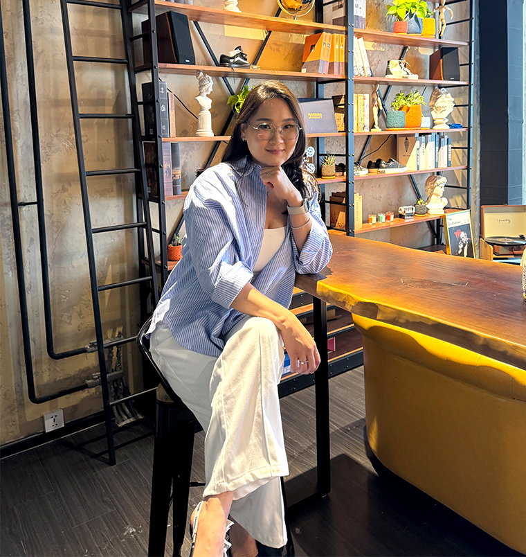 A person sits on a stool by a wooden table in a stylish workspace, wearing a striped shirt and glasses. Behind them is a shelf with books and decorative items. The space has a modern, cozy vibe.