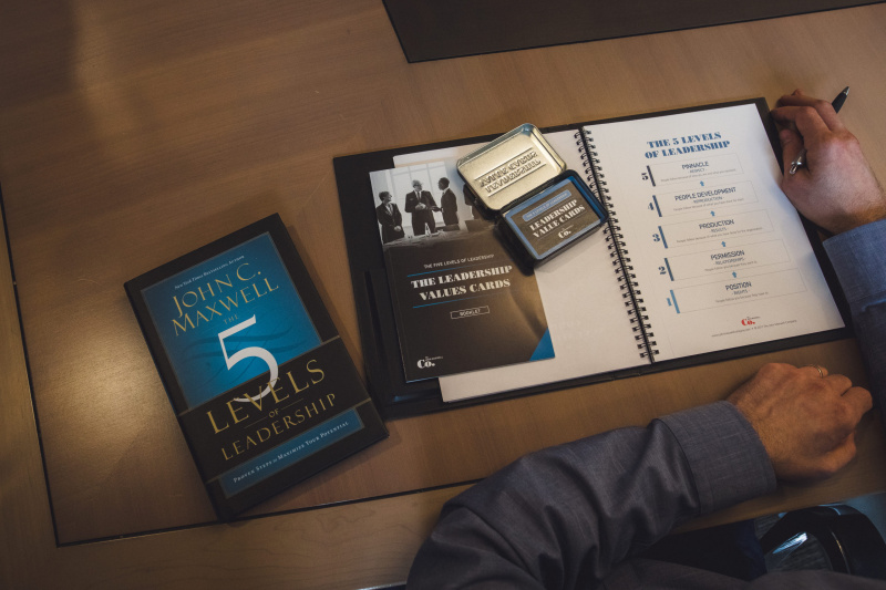 A person writes on a paper titled "The 5 Levels of Leadership." Nearby are a book called "5 Levels of Leadership" by John C. Maxwell and a tin labeled "The Leadership Values Cards." They're sitting at a wooden table.